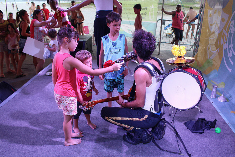 Crianças no palco com um homem com um violão e um tambor nas costas