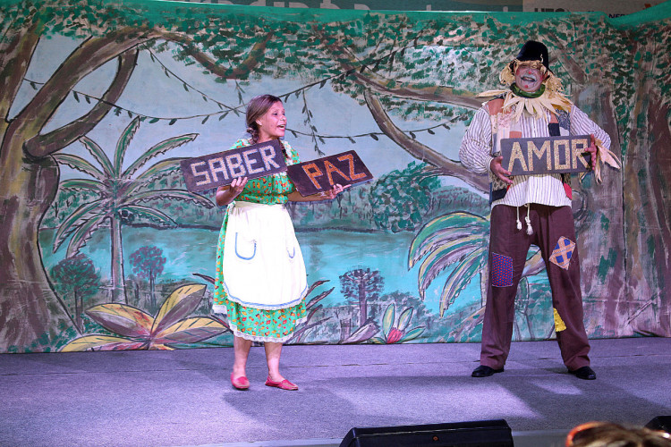 Atores seguram placas com os dizeres "saber", "paz" e "amor" em um palco