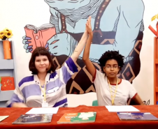 Duas mulheres atrás de uma mesa com uma ilustração ao fundo