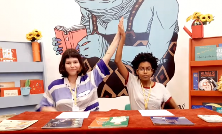 Duas mulheres atrás de uma mesa com uma ilustração ao fundo