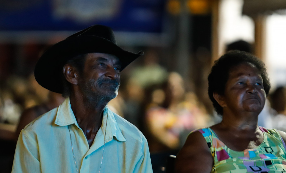 Um homem de chapéu preto e camisa clara assistindo o cinema junto de uma mulher de vestido colorido