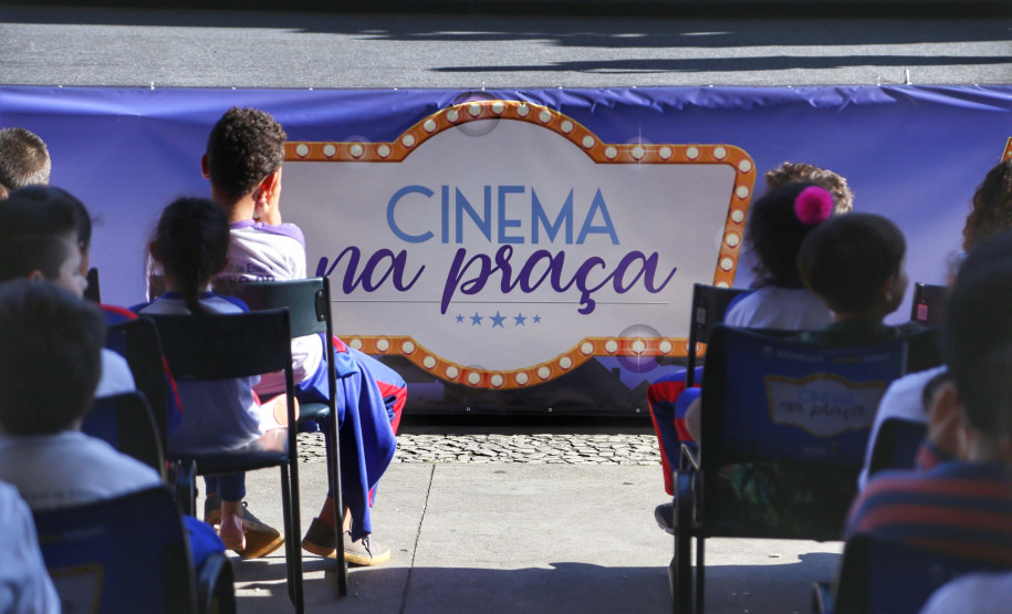 Crianças sentadas com o banner do projeto Cinema na Praça logo a frente